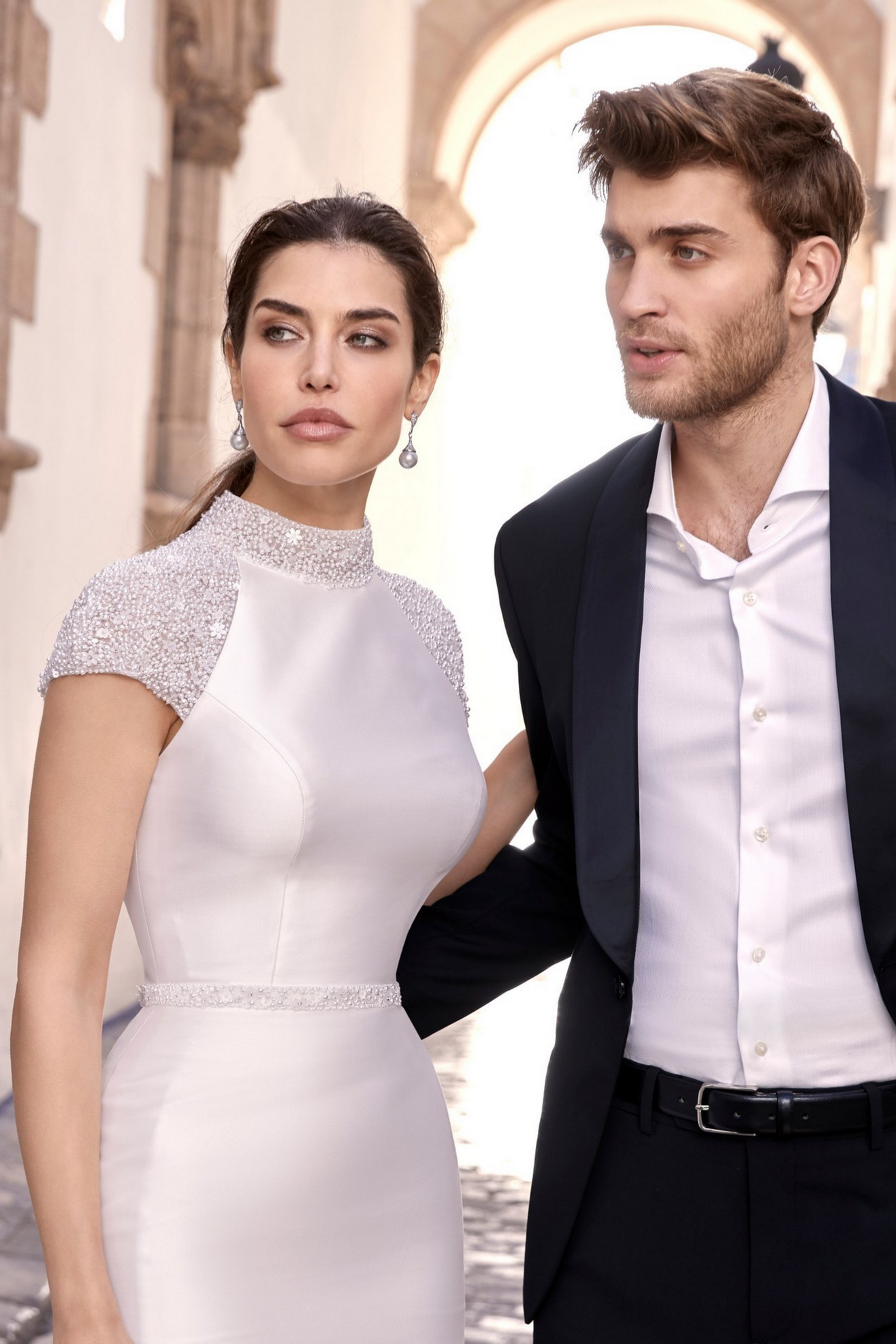 Man with arm around bride wearing fitted mikado wedding dress with beaded high neckline, cap sleeves and belt detail